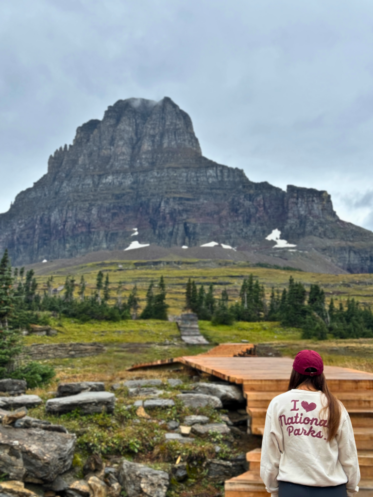 "Girl standing at Glacier National Park, overlooking a mountainous landscape while wearing a sweatshirt with 'I love National Parks' written in bold red letters on the back."