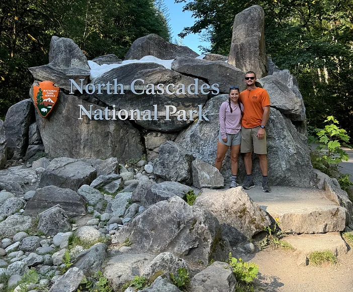 A couple posing with the North Cascades National Park entrance sign, symbolizing the start of their adventure and successful Planning a National Park Trip.