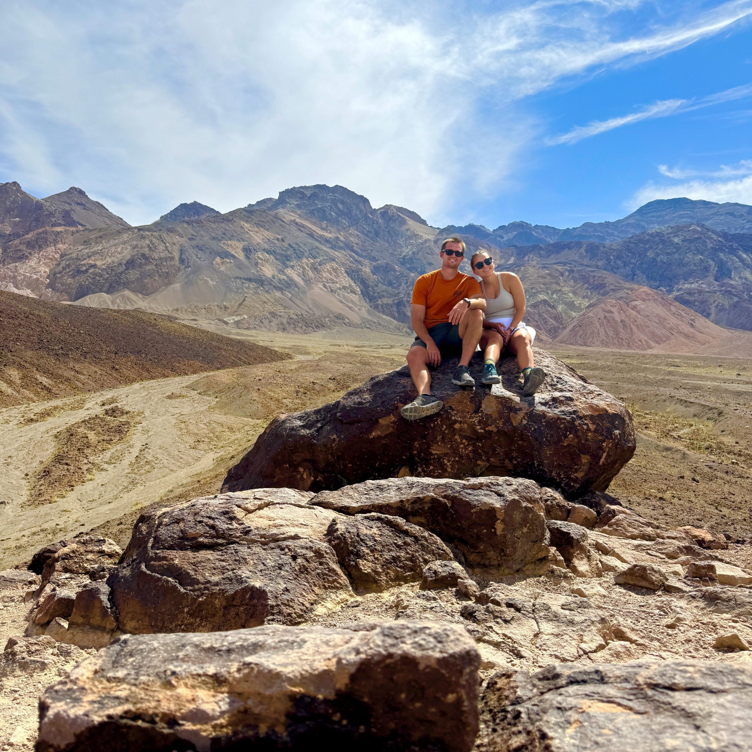 A couple posing on a rock with the vibrant, multicolored mountains of Artists Drive in Death Valley National Park in the background, showcasing the park's stunning desert landscape.