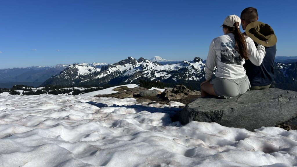 Two hikers sitting on a rock at snowy Mount Rainier National Park, gazing at distant volcanoes under a clear sky, embodying the spirit of an outdoor travel blog.