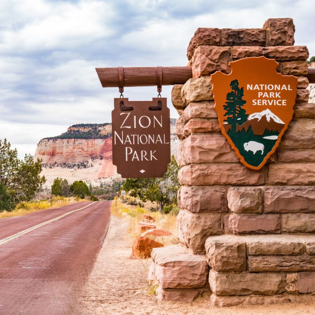 Zion National Park entrance sign surrounded by red rock formations and greenery, a must-see for those exploring the best time to visit US National Parks.