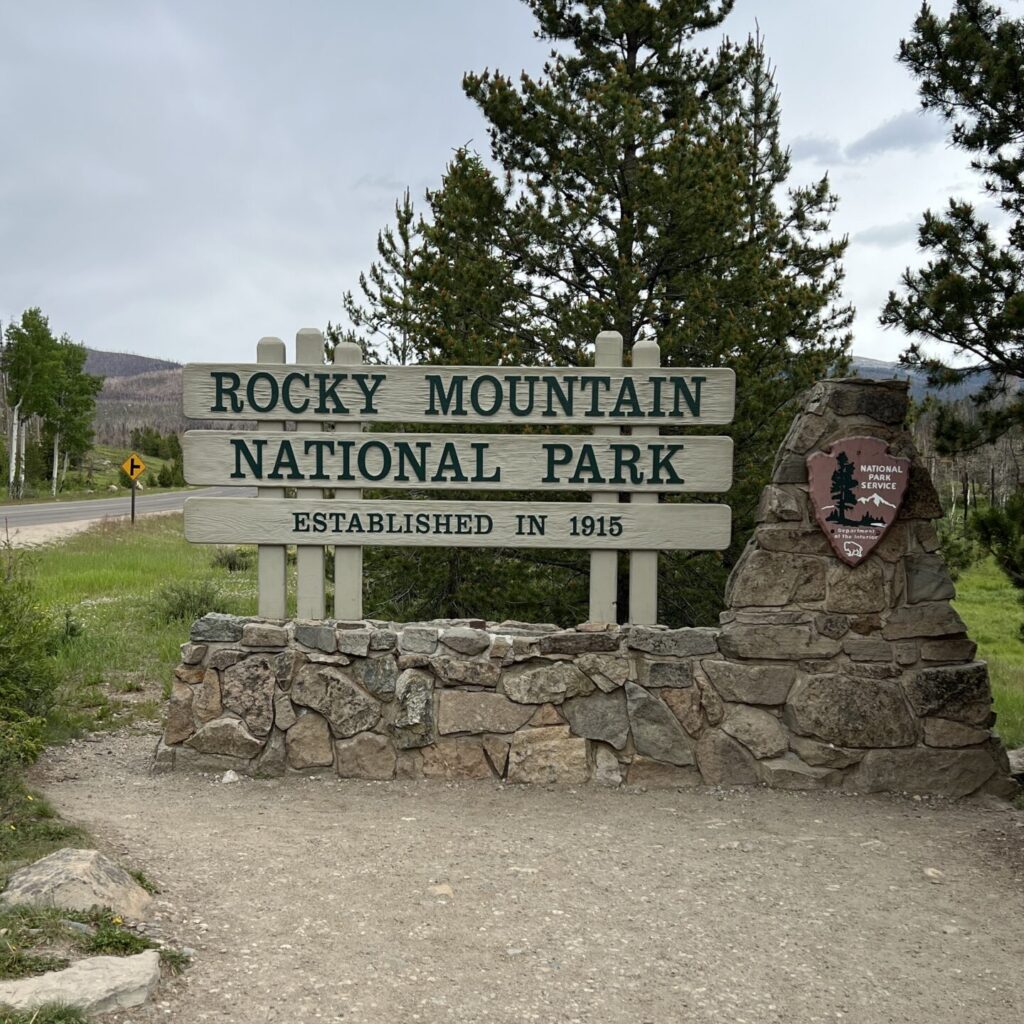 Rocky Mountain National Park Entrance Sign – A wooden entrance sign for Rocky Mountain National Park, surrounded by tall pine trees and set against a mountainous backdrop.